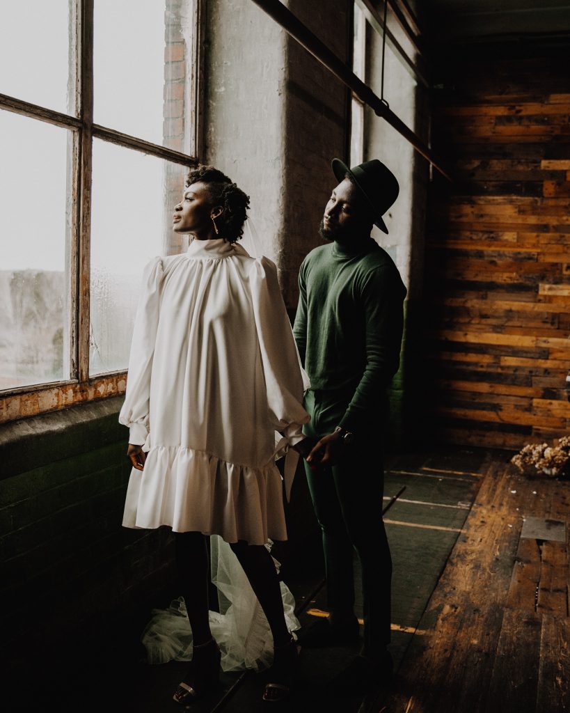 Newly married couple stand together looking out of a window. The bride wears a short Moonchild wedding dress with long sleeves and a veil