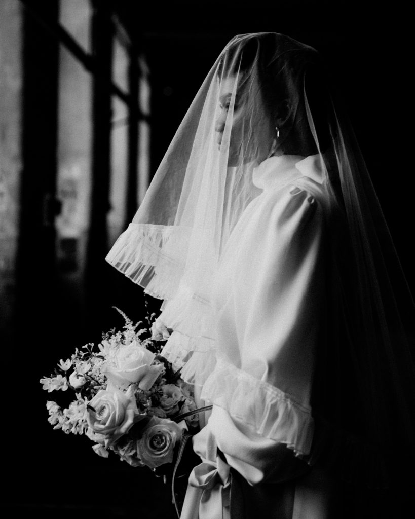 Black and white image of newlywed bride wearing a short Moonchild wedding dress with a veil over her face carrying a wedding bouquet