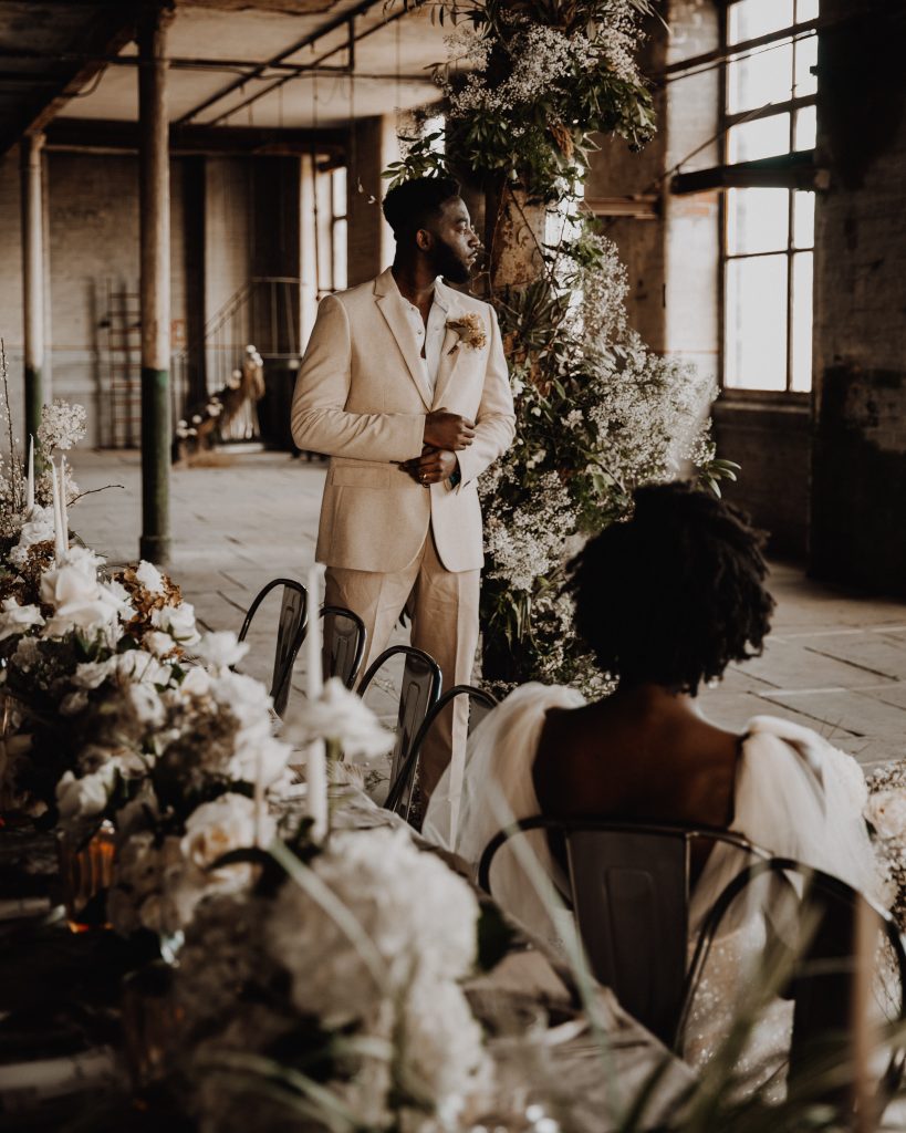 Bride sits at her wedding table whilst her newly married groom stands next to her at their wedding venue