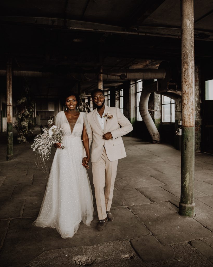 Bride and groom walk hand in hand towards the camera. She wears a Berta wedding gown that sparkles and carries a white wedding bouquet