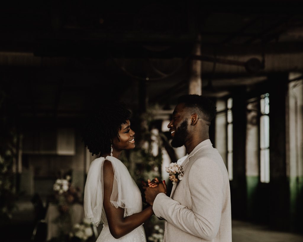 Newly married wedding couple gaze at each other holding hands. The bride wears a sparkly Berta wedding gown