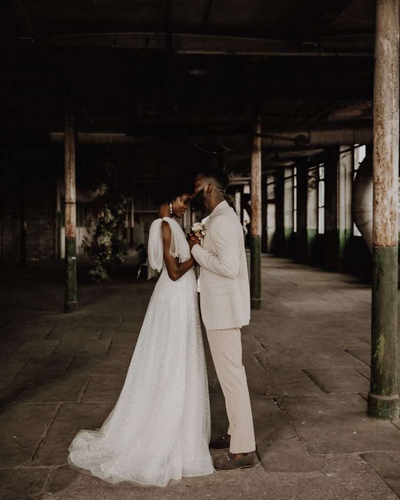 A portrait image of a bride and groom on their wedding day holding hands. The groom wears a tan suit and the bride wears a long flowy Berta wedding dress