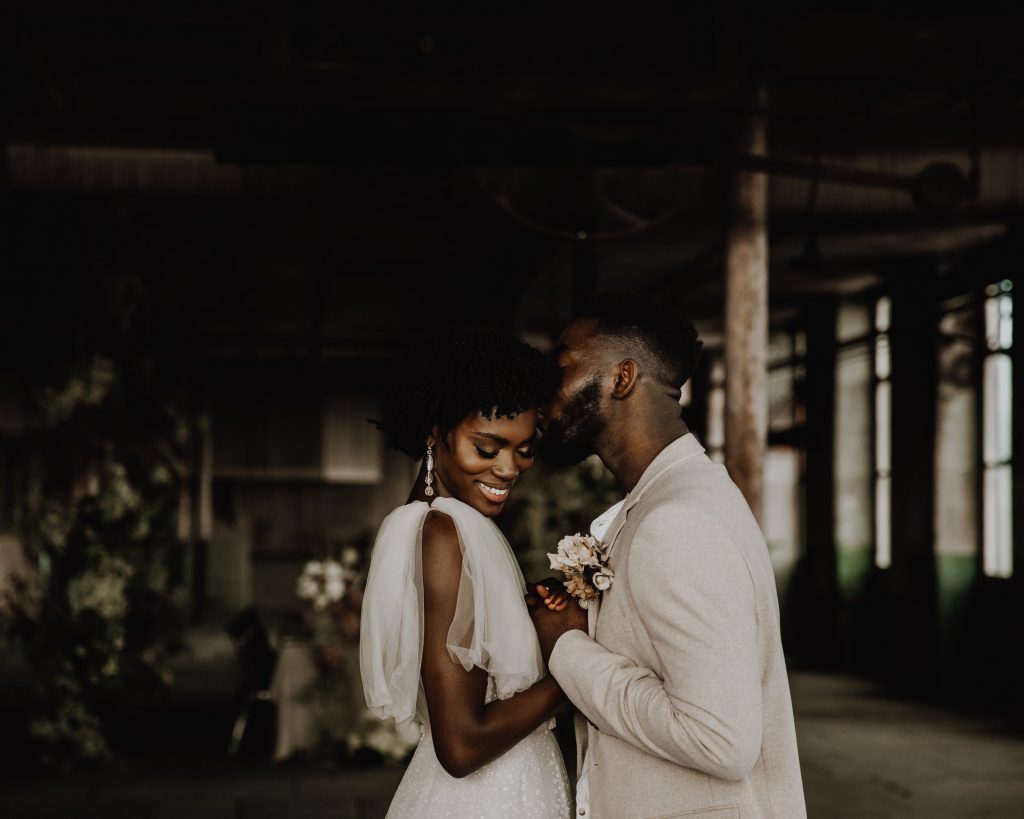 Newlywed couple embracing. The groom kisses his bride gently on the forehead. She wears a Berta wedding gown