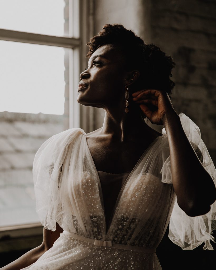 A bride looking thoughtful on her wedding day. She wears a plunging v neck sparkling Berta wedding gown with tulle shoulder ties