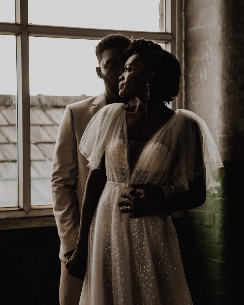 A bride and groom stand close together on their wedding day. She wears a plunging v neck Berta wedding gwon with head to toe sparkle
