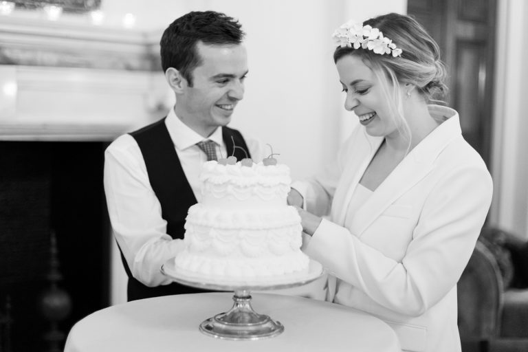 Cutting of the two tier cream wedding cake with piped detailing and charry topping for low key summer micro wedding