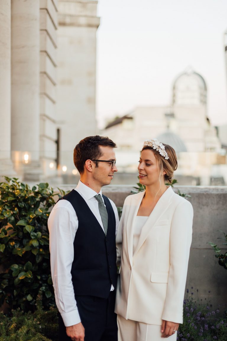 Bride and groom stand together during rooftop city micro wedding as bride wears white blazer and flower crown