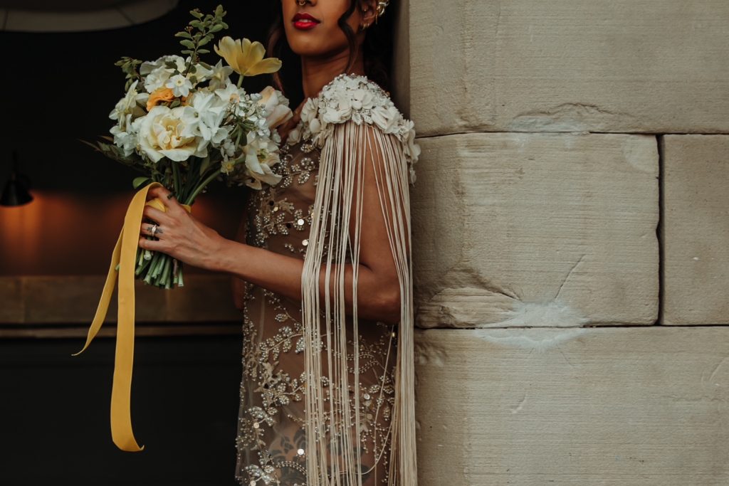 A bride leans against a stone wall holding her wedding bouquet