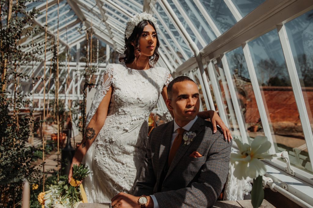 A groom sits down in a Victorian greenhouse whilst his bride wearing a white wedding dress stands behind him