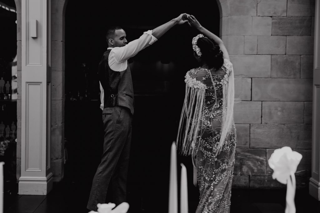 Black and white image of a bride and groom having their first dance on their wedding evening. The bride wears a heavily embellished wedding dress with detailed fringing