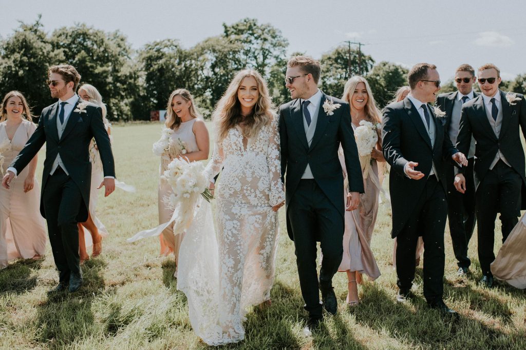 Bride and groom relax in fields with wedding party during summer celebrations