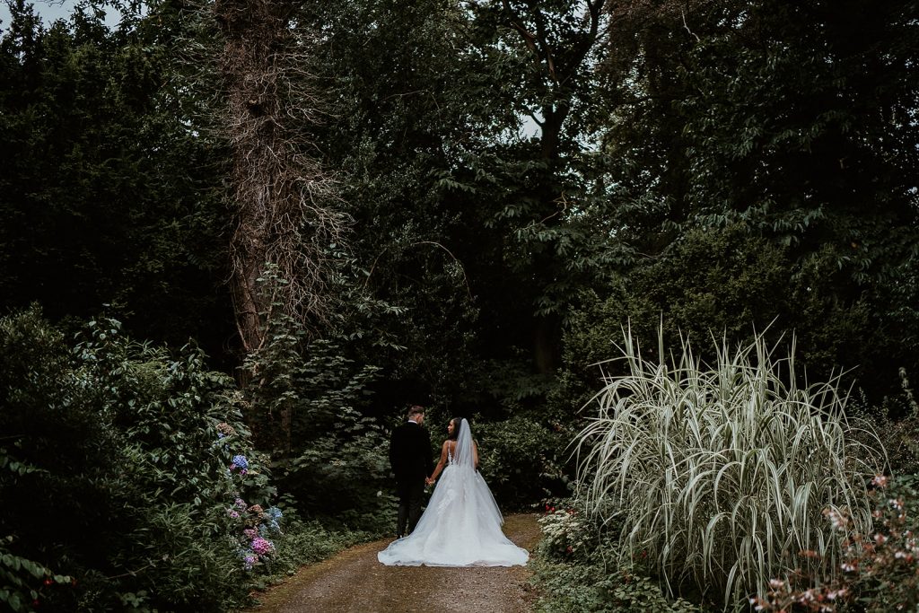 Bride and groom stand with their back to the camera surrounded by lush greenery and trees during a quiet couple moment on their wedding day