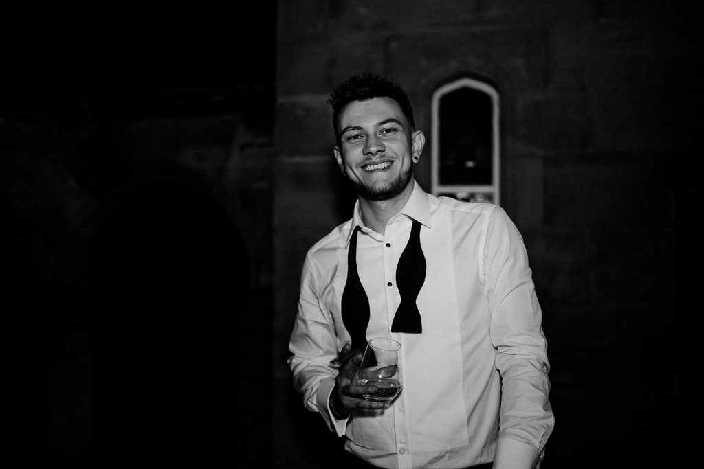 A groom looking relaxed towards the end of his wedding evening, with his bow tie undone, holding a drink