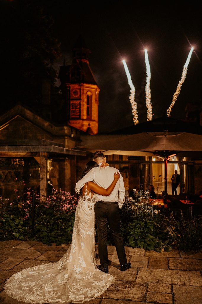 A bride and groom stand holding each other on the terrace of their wedding venue watching fireworks