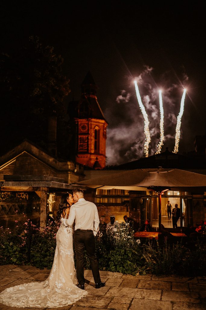 A bride and groom stand holding each other on the terrace of their wedding venue watching fireworks