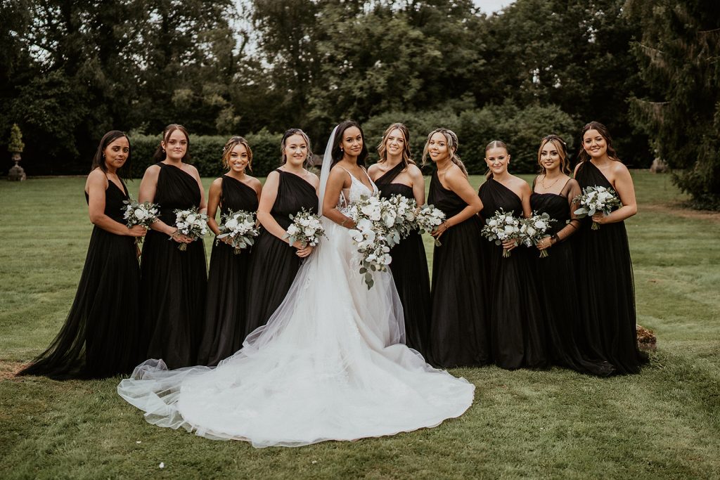 Bride stands in the centre of the photo wearing a large white Enzoani wedding dress surrounded by her bridesmaids who wear black bridesmaids dresses