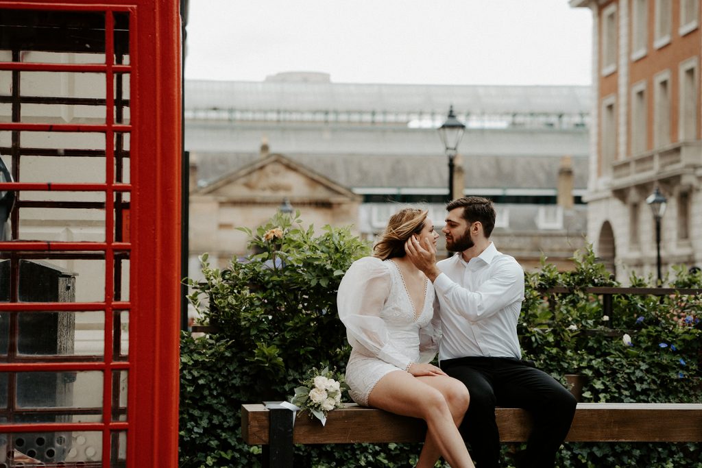 A groom tenderly strokes his wife's face as they sit on a bench with her wedding bouquet next to her