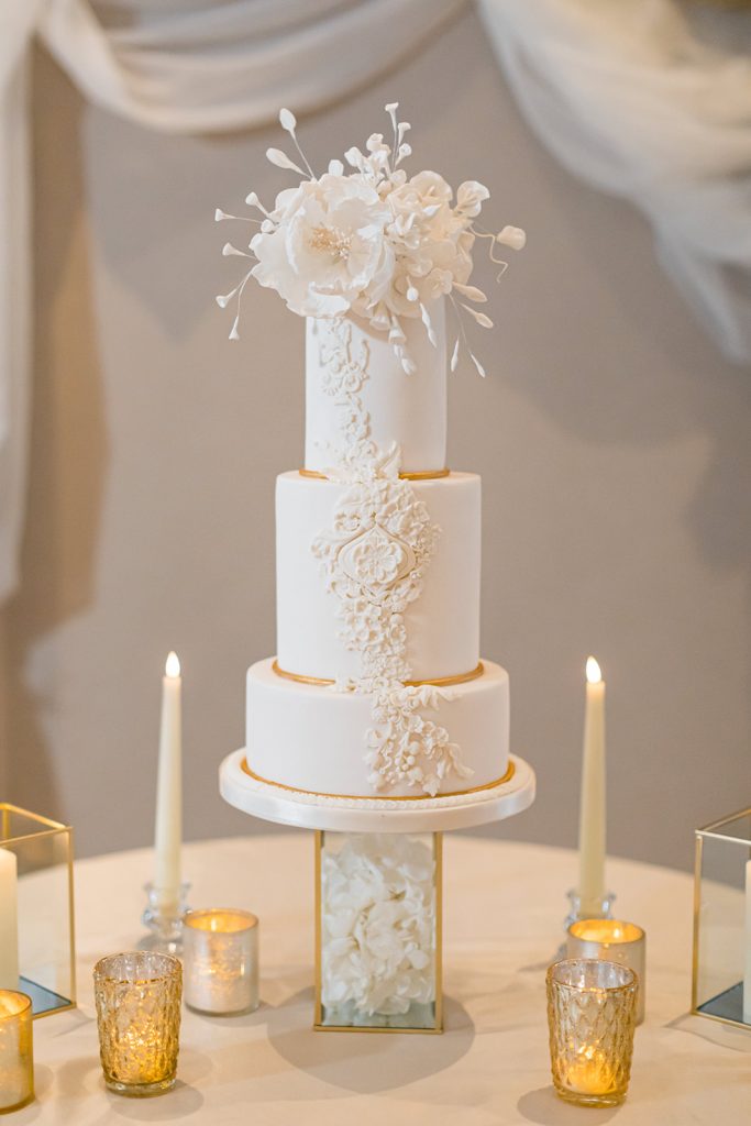 A three tiered white wedding cake with intricate piped detailing and a hint of gold