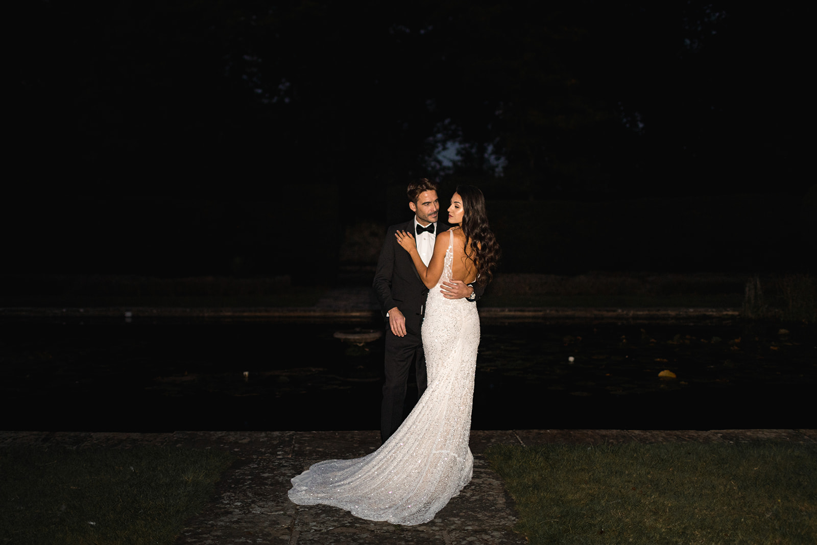 A black tie wedding couple snuggle up to each other outside underneath a dusky twilight blue sky