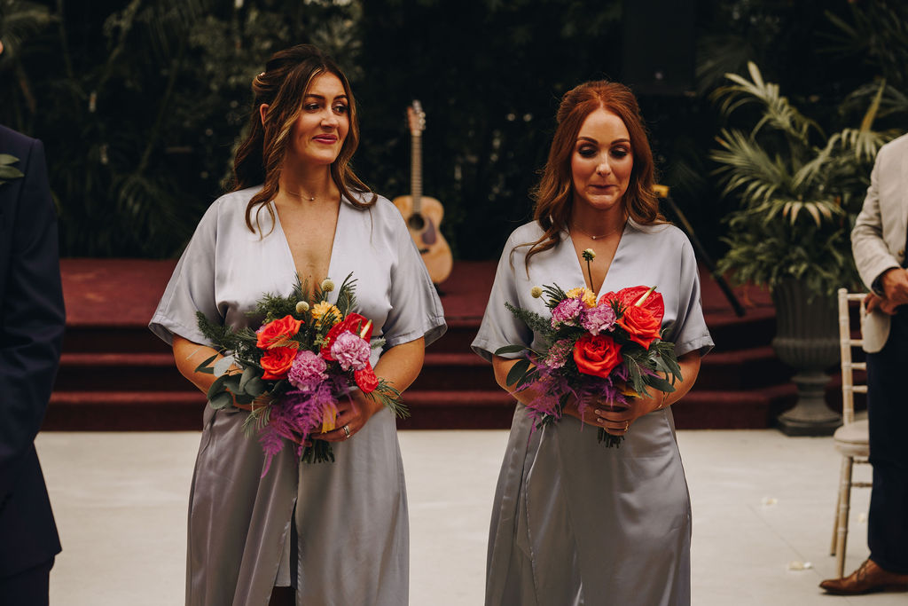 Bridesmaids wearing matching satin V-neck silvery blue bridesmaids dresses with a colourful wedding bouquet walking up the aisle