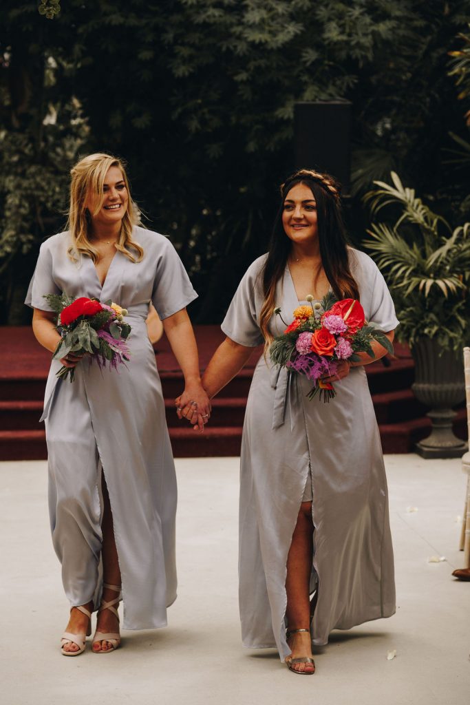 Bridesmaids wearing matching satin V-neck silvery blue bridesmaids dresses with a colourful wedding bouquet walking up the aisle