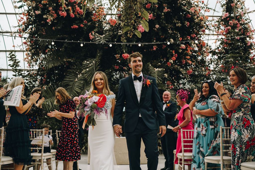 A bride and groom walk back down the aisle having now officially been married. Their loved ones stand and clap as they walk down the aisle as husband and wife