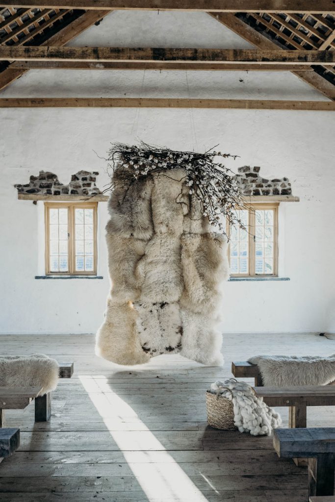 A sheepskin wedding ceremony backdrop styled with twigs, cotton and blossom for a modern barn wedding
