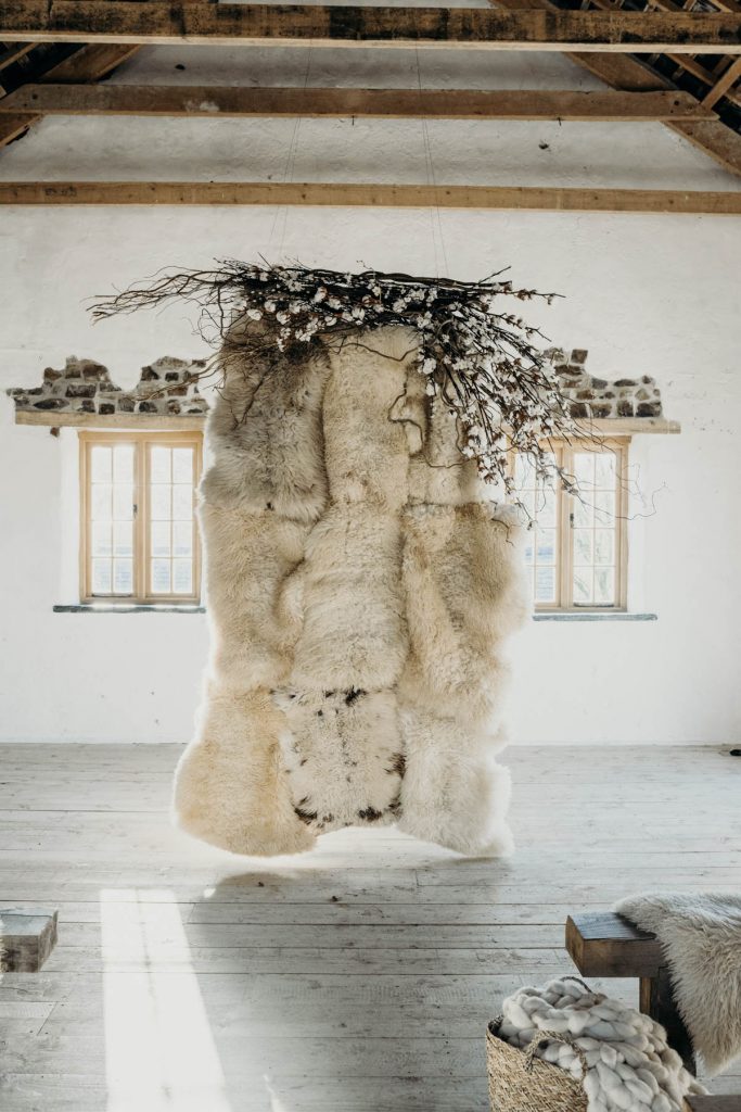 A sheepskin wedding ceremony backdrop styled with twigs, cotton and blossom for a modern barn wedding