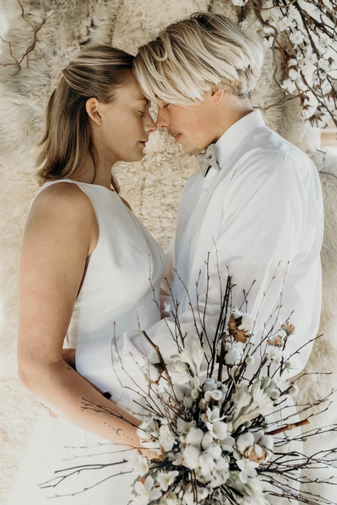 A modern bride and groom gently press foreheads together whilst the bride holds a bouquet of twigs, blossom and cotton