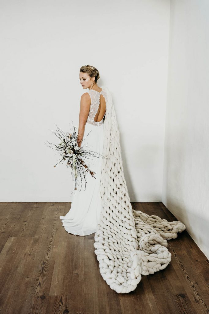 A modern barn bride in a white wedding gown, cosy in a thick cable knit woollen blanket