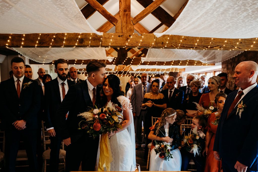 A grrom gently kisses his wife to be on her forehead during their wedding ceremony, surrounded by their loved ones behind them