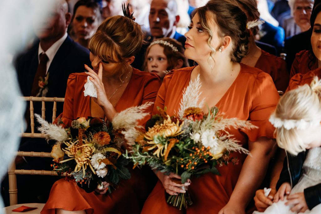 A bridesmaid in a burnt orange bridesmaids dress wipes a tear from her eye during an emotional wedding ceremony