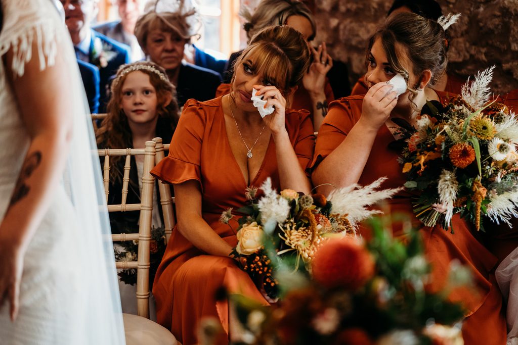 A bridesmaid in a burnt orange bridesmaids dress wipes a tear from her eye during an emotional wedding ceremony