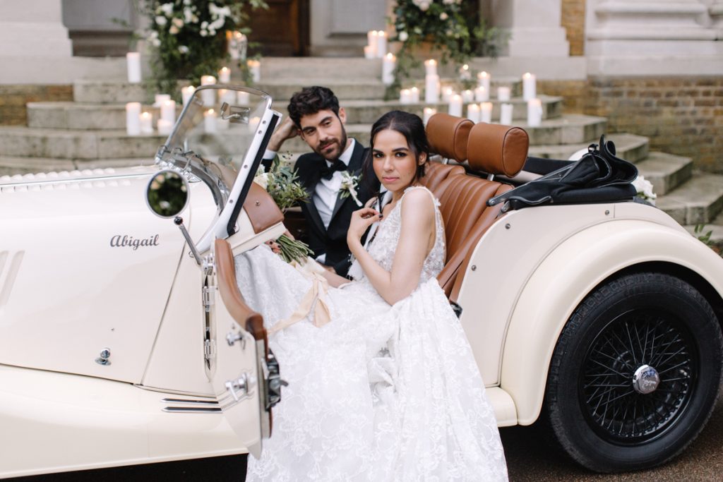 An elegant bride and groom sit in their convertible wedding car with a backdrop of pillar candles lining the stairs to their wedding venue