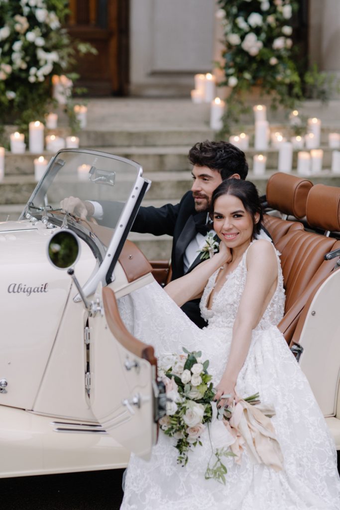 An elegant bride and groom sit in their convertible wedding car with a backdrop of pillar candles lining the stairs to their wedding venue