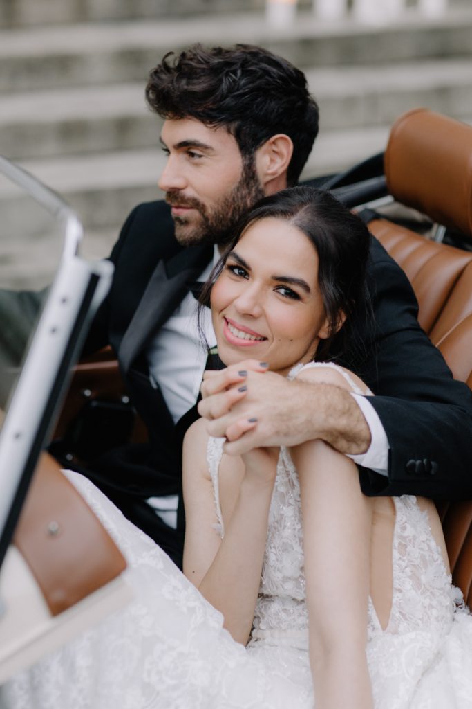 An elegant bride and groom sit in their convertible wedding car with a backdrop of pillar candles lining the stairs to their wedding venue