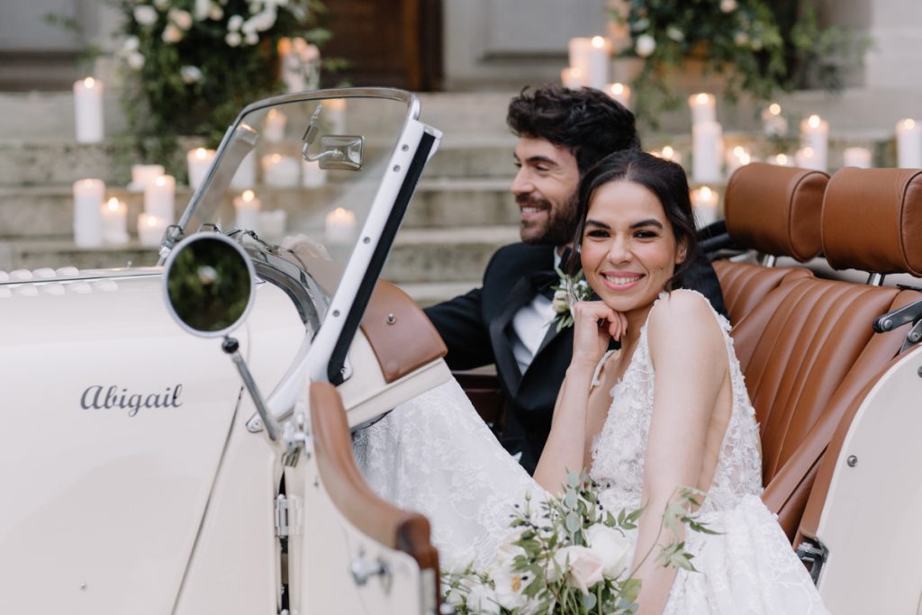 An elegant bride and groom sit in their convertible wedding car with a backdrop of pillar candles lining the stairs to their wedding venue