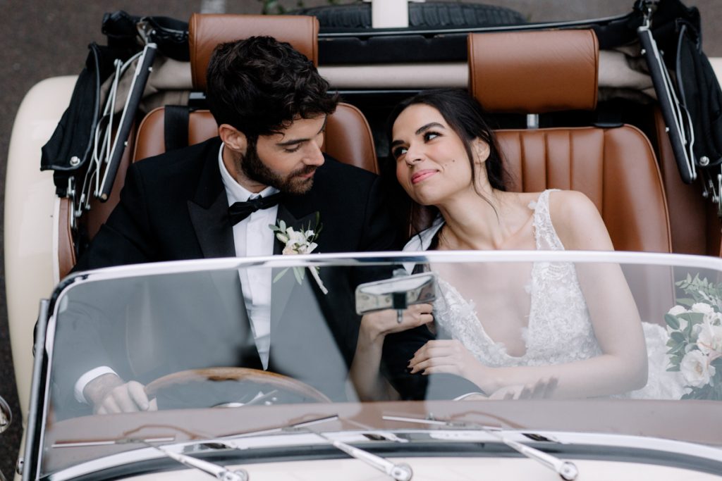 An elegant bride and groom sit in their convertible wedding car with a backdrop of pillar candles lining the stairs to their wedding venue