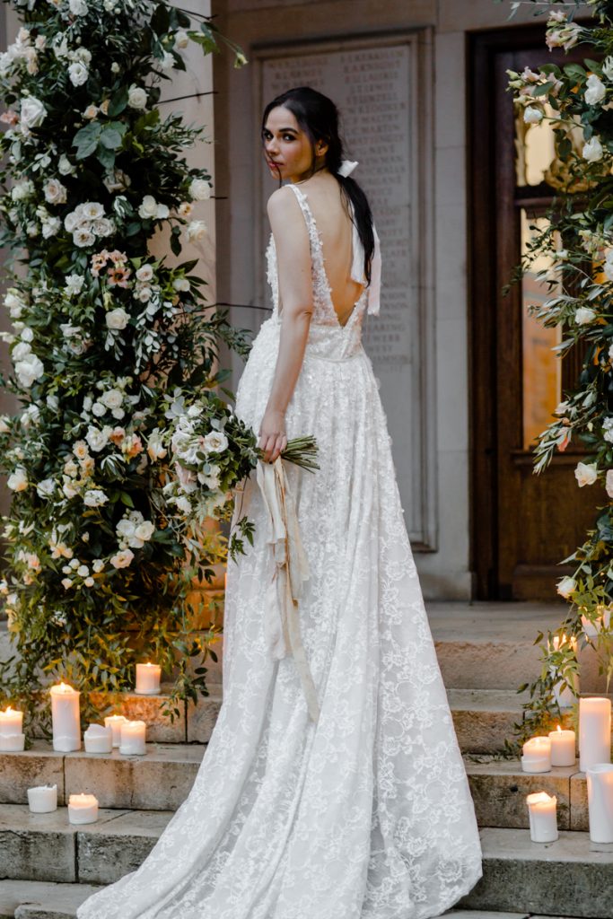 A bride wearing an Inbal Dror wedding gown stands on the outdoor steps to her wedding venue which are styled with masses of pillar candles and framed by two columns covered in pink and white flowers