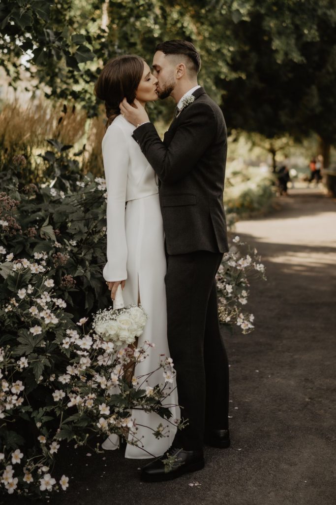 Two newlyweds share a gentle kiss just after their wedding ceremony, with a backdrop of natural flowers in the city of Bath