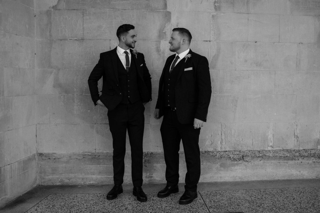 A groom stands with his best man on his wedding day. Both gentlemen look smart in their suits and ties