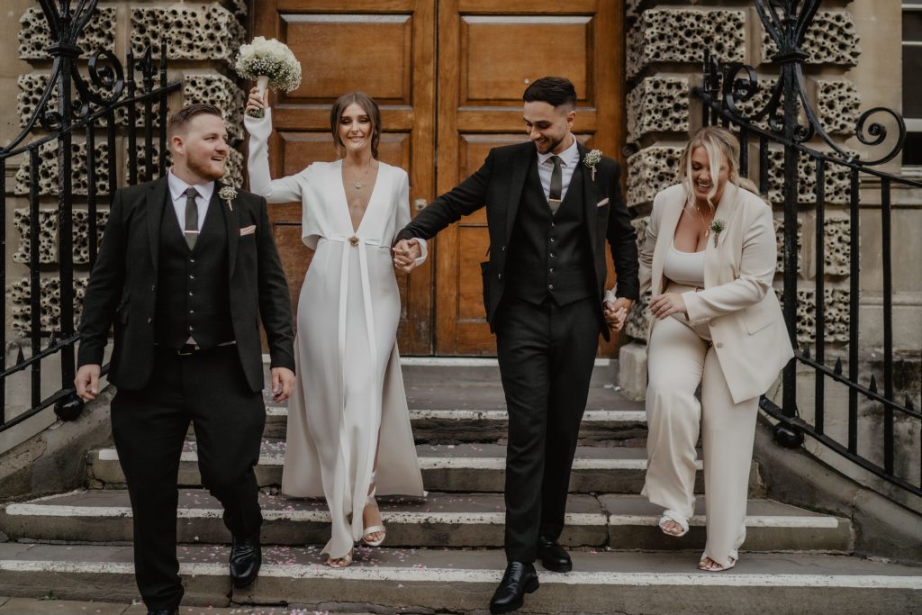 A newlywed couple stand on the stairs to their city ceremony venue with their two best friends during their elopement