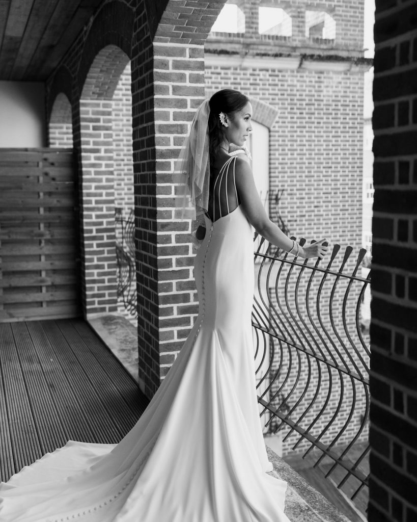 A black and white portrait of a bride wearing a fitted wedidng dress with intricate back detailing and a short wedding veil. She wears a full silver embellished ear cuff in one ear