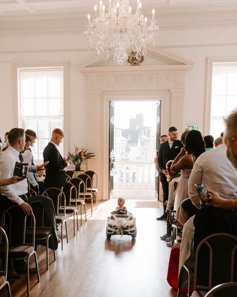 The bride and grooms baby daughter 'drives' down the ceremony aisle in a white Mercedes electric toy car with white wedding ribbon on the bonnet. She wears a cute white outfit and white headband. The guests in the aisles look with delight and joy as she 'drives', bearing the wedding rings