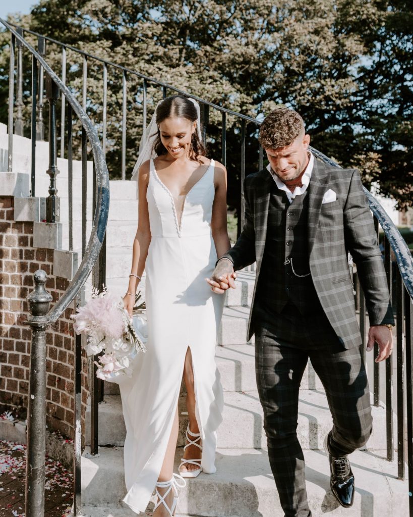 A bride and groom walk and hold hands. The froom wears a checked 3 piece wedding suit and a small button hole. The bride wers a fitted wedding dress with a thigh high split and holds a small wedding bouquet of white and pink flowers