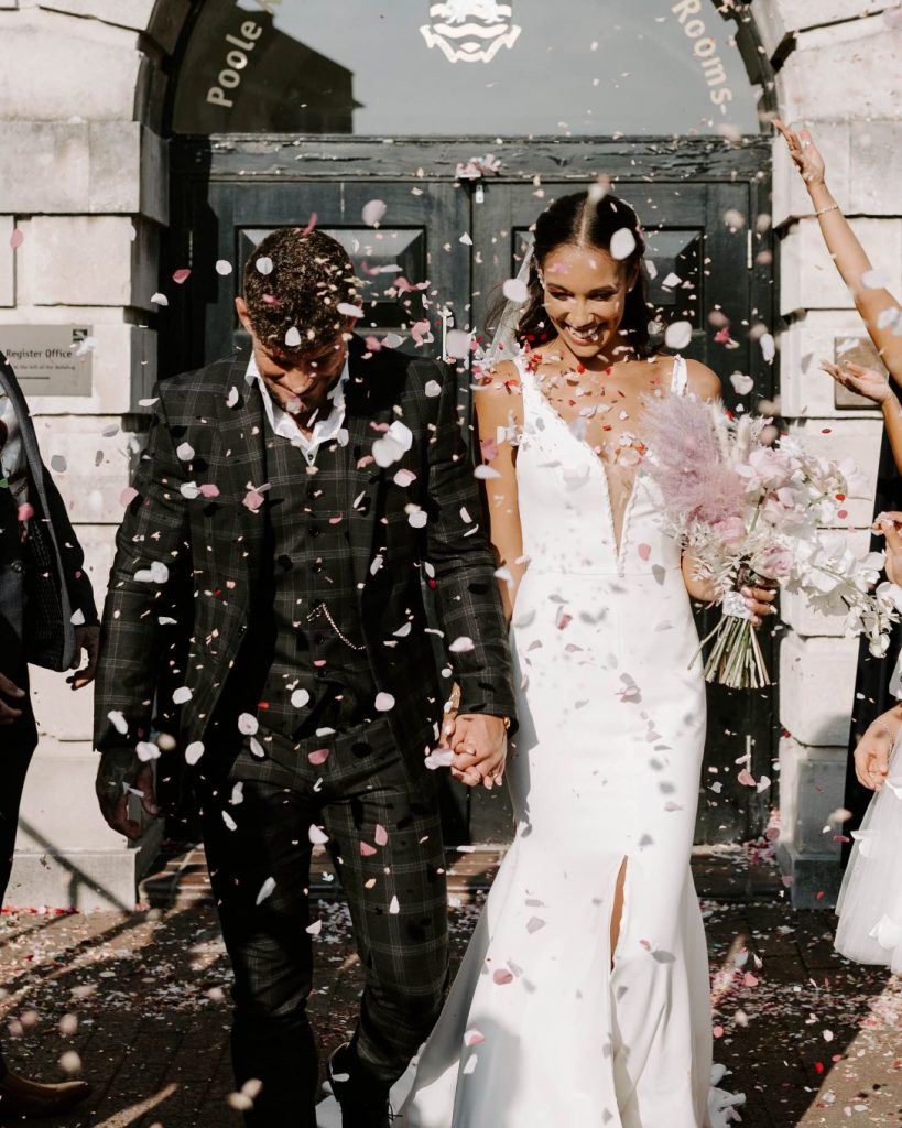A newly married bride and groom walk outside in the sunshine after their wedding ceremony as they are showered with pink and white confetti