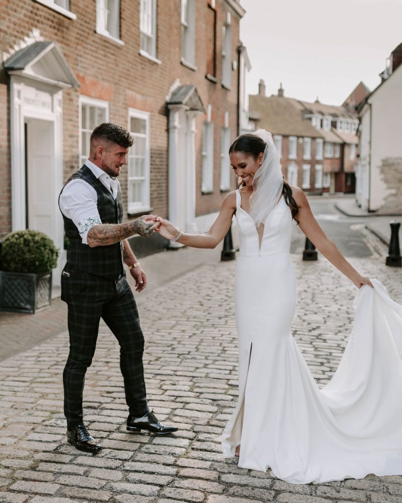 A bride and groom have a private dance on a quiet street. The groom wears a waistcoat with a shirt rolled up at the sleeves as he gently holds his wife's hand and spins her around. The bride wears a fitted white wedding dress with a short veil