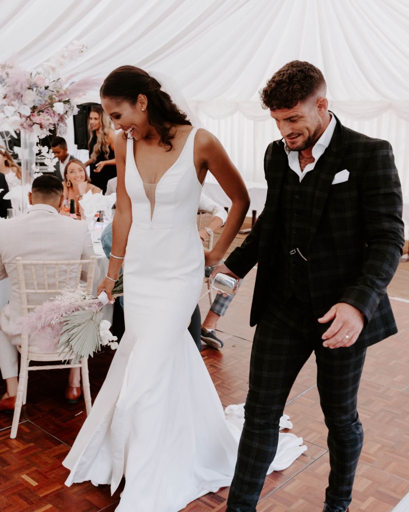 A newly married bride and groom walk hand in hand into their wedding breakfast room where their guests are sat on round table.s The couple smile as they look down at the floor
