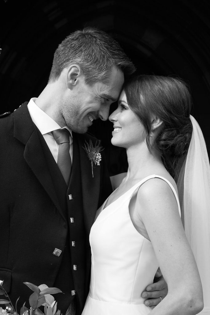 A black and white couple portrait of a newly married bride and groom, gazing at one another and smiling. The bride holds a large wedding bouquet by her waist