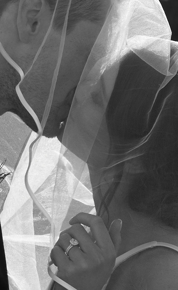 A black and white image of a bride and groom kissing tenderly underneath the brides veil. You can see a large, beautiful diamond on the brides hand